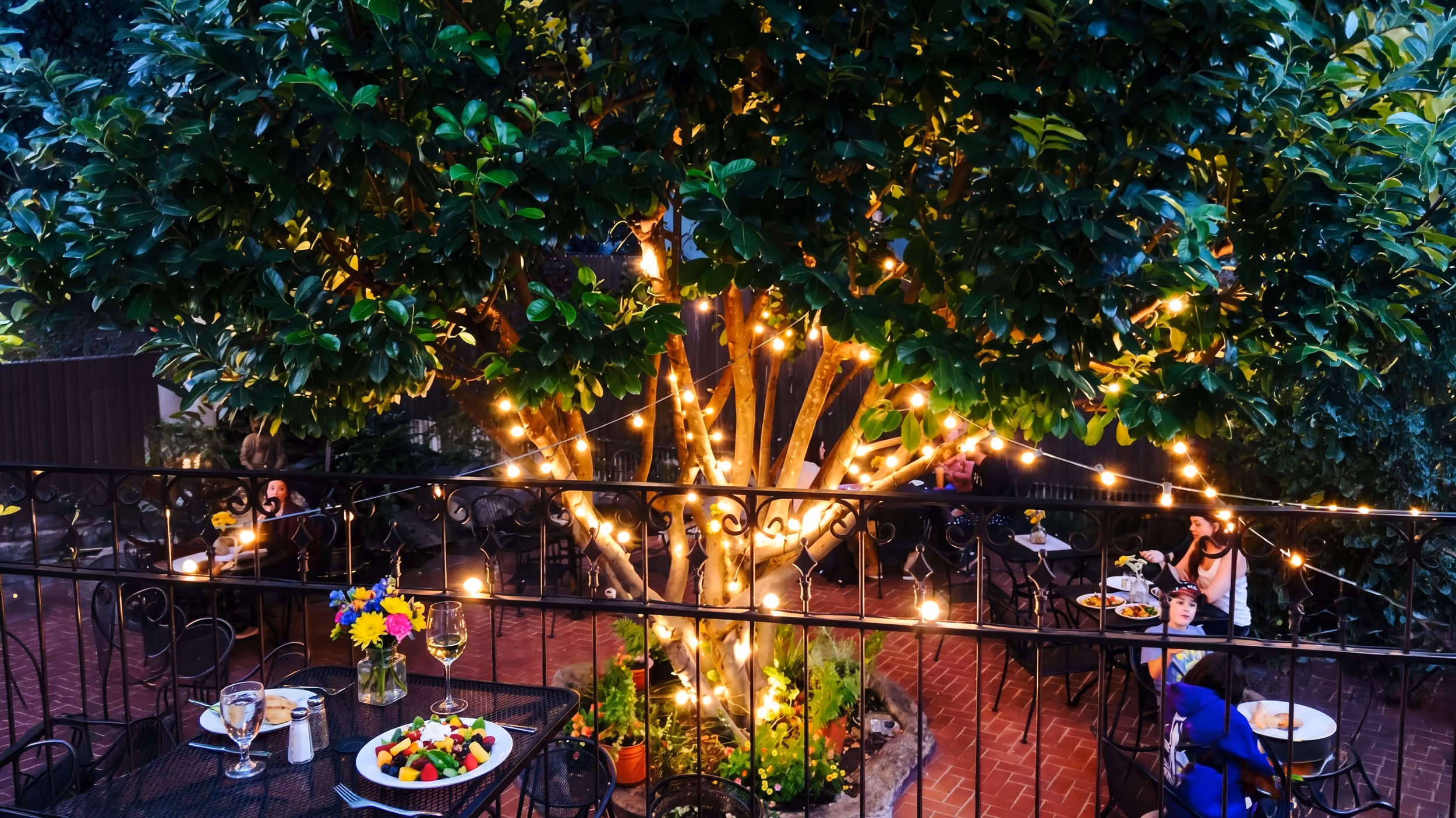Outdoor patio dining area with string lights wrapped around a large tree and people eating at tables.
