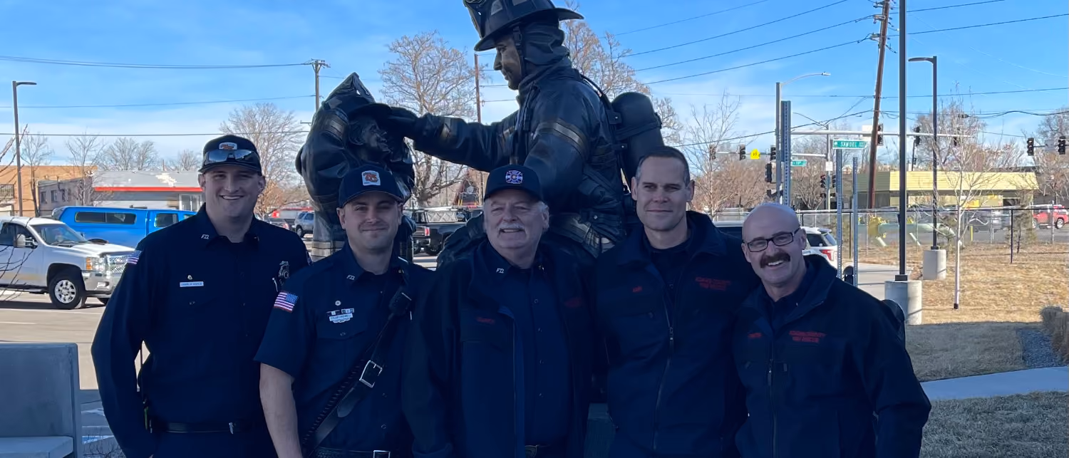 Five firefighters in uniform posing outdoors in front of a firefighter statue on a clear day.