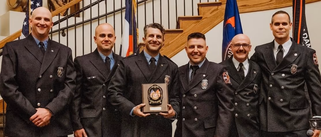 Six men in formal firefighter uniforms standing in front of a staircase, one holding an award plaque.