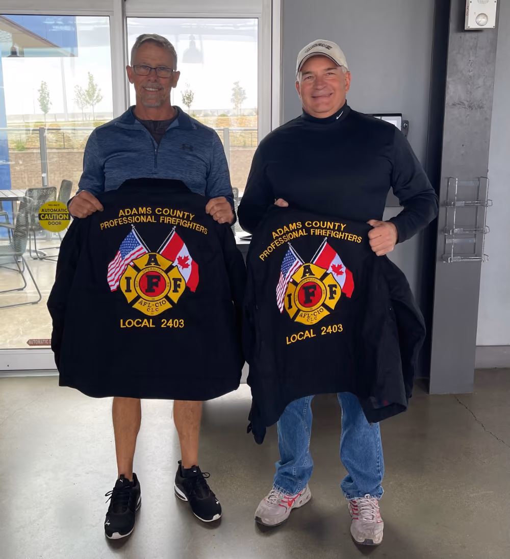Two men holding jackets with Adams County Professional Firefighters Local 2403 logo featuring US and Canadian flags.