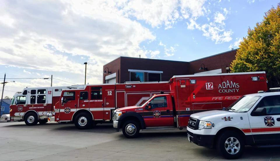 Three Adams County fire rescue vehicles parked near a brick fire station under a partly cloudy sky.