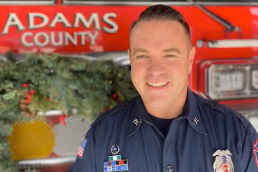 Smiling firefighter in uniform standing in front of Adams County fire truck with wreath decoration.