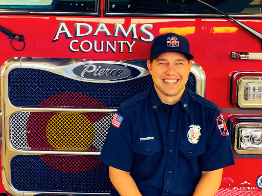 A firefighter in uniform smiling and standing in front of a red Adams County fire truck.
