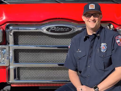 Smiling firefighter in uniform and cap stands in front of a red fire truck grille with Pierce logo.