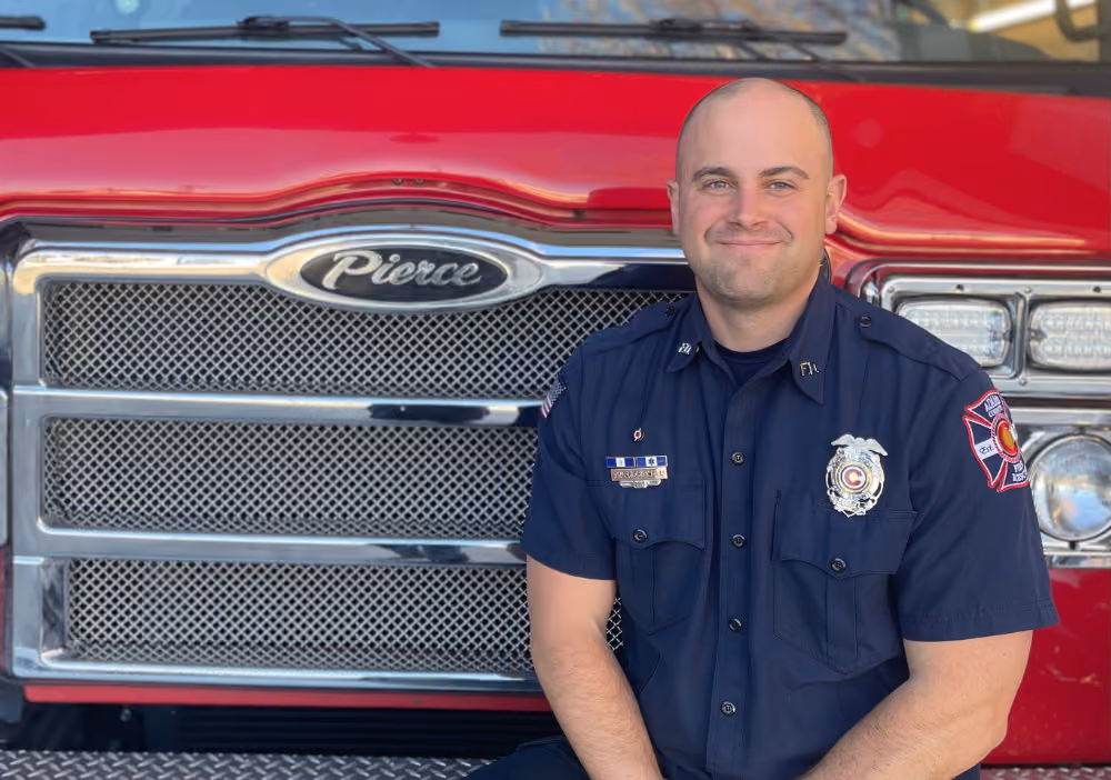 Smiling firefighter in uniform sitting in front of a red Pierce fire truck.