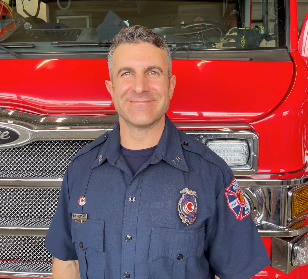 Smiling firefighter in uniform standing in front of a red fire truck.