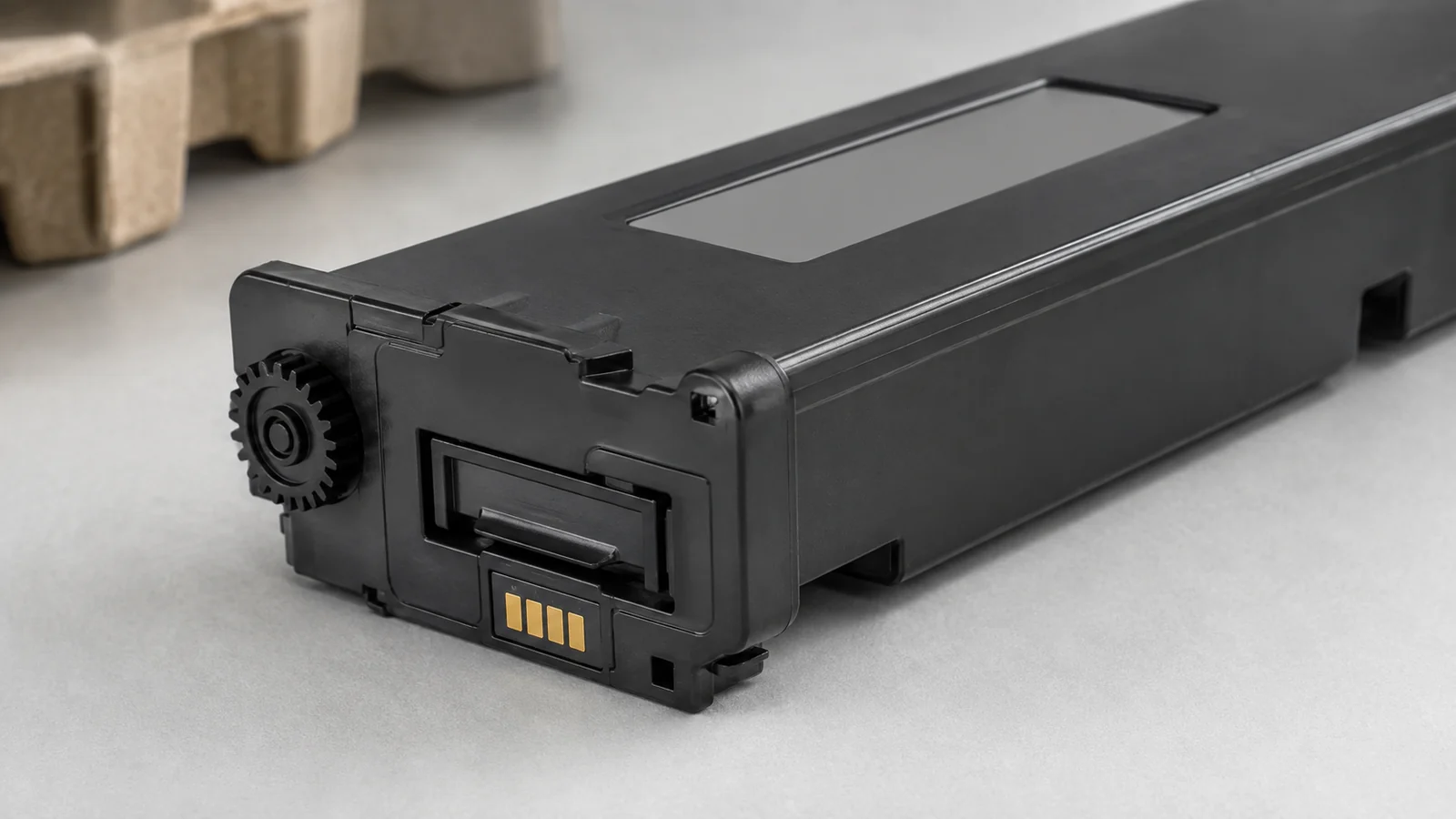 Detalhe de um recipiente genérico de toner com área de etiqueta em branco, guias moldadas, saída fechada e pequenos contatos dourados.