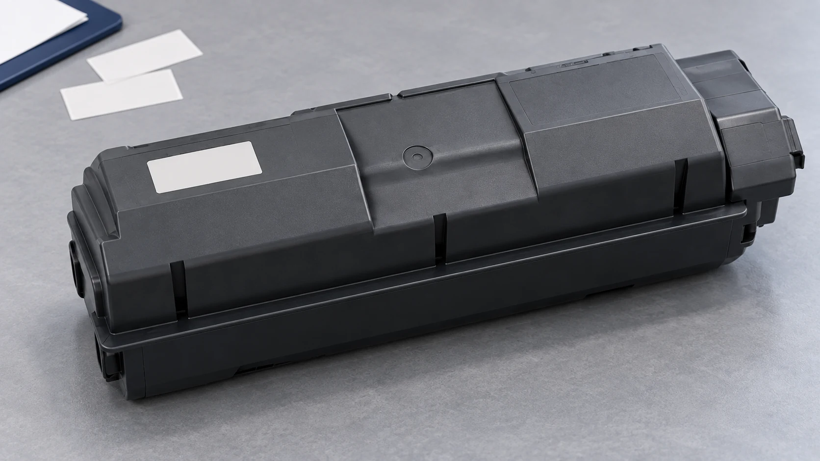 Close view of a closed black toner supply container with a blank label and molded locating edges on a gray inspection surface.