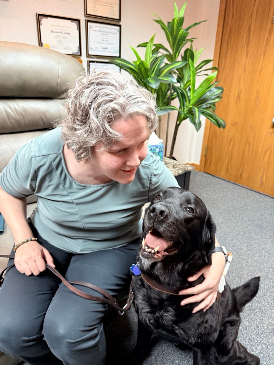 Beth Gustin petting Manolo, a happy black dog, in office with plants and diplomas