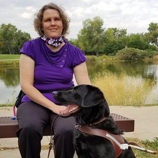 Beth Gustin, in purple shirt sitting with black dog, Manolo, in a park with a lake