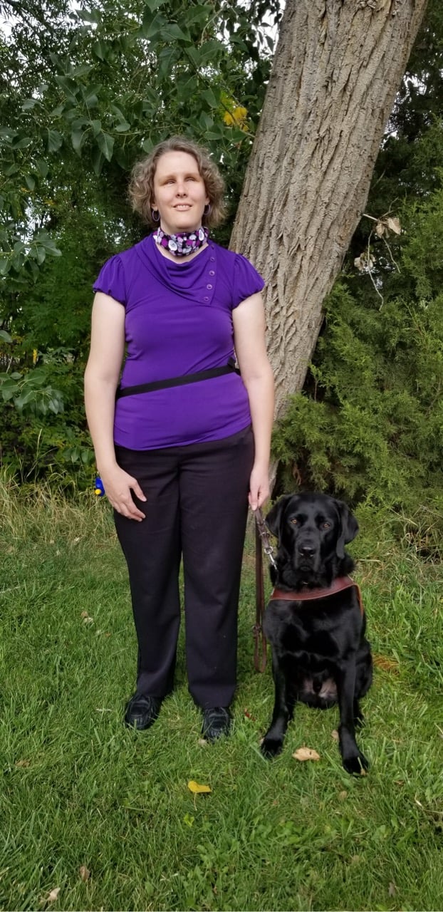 Beth Gustin, in purple shirt standing with black Labrador dog, Manolo, under large tree outdoors