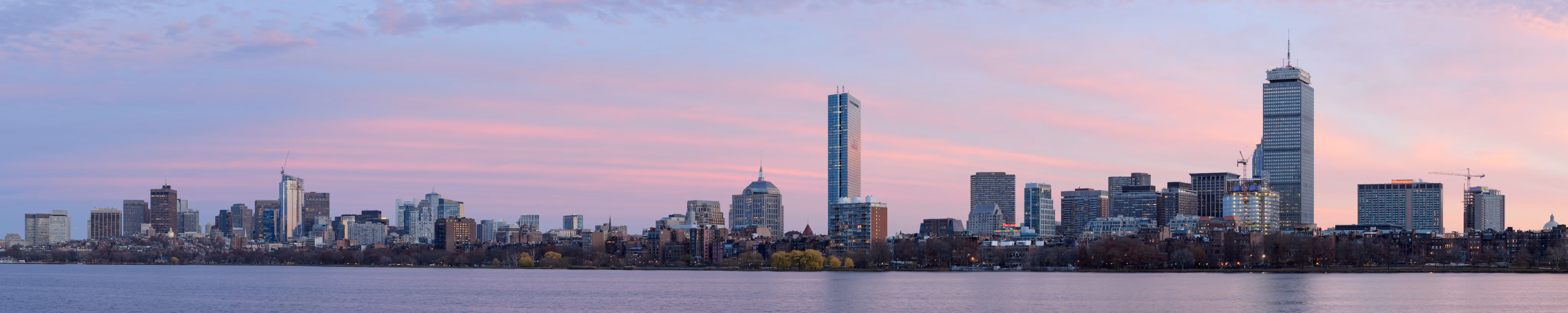 Panoramic view of a city skyline at sunset with pink clouds over calm river water.
