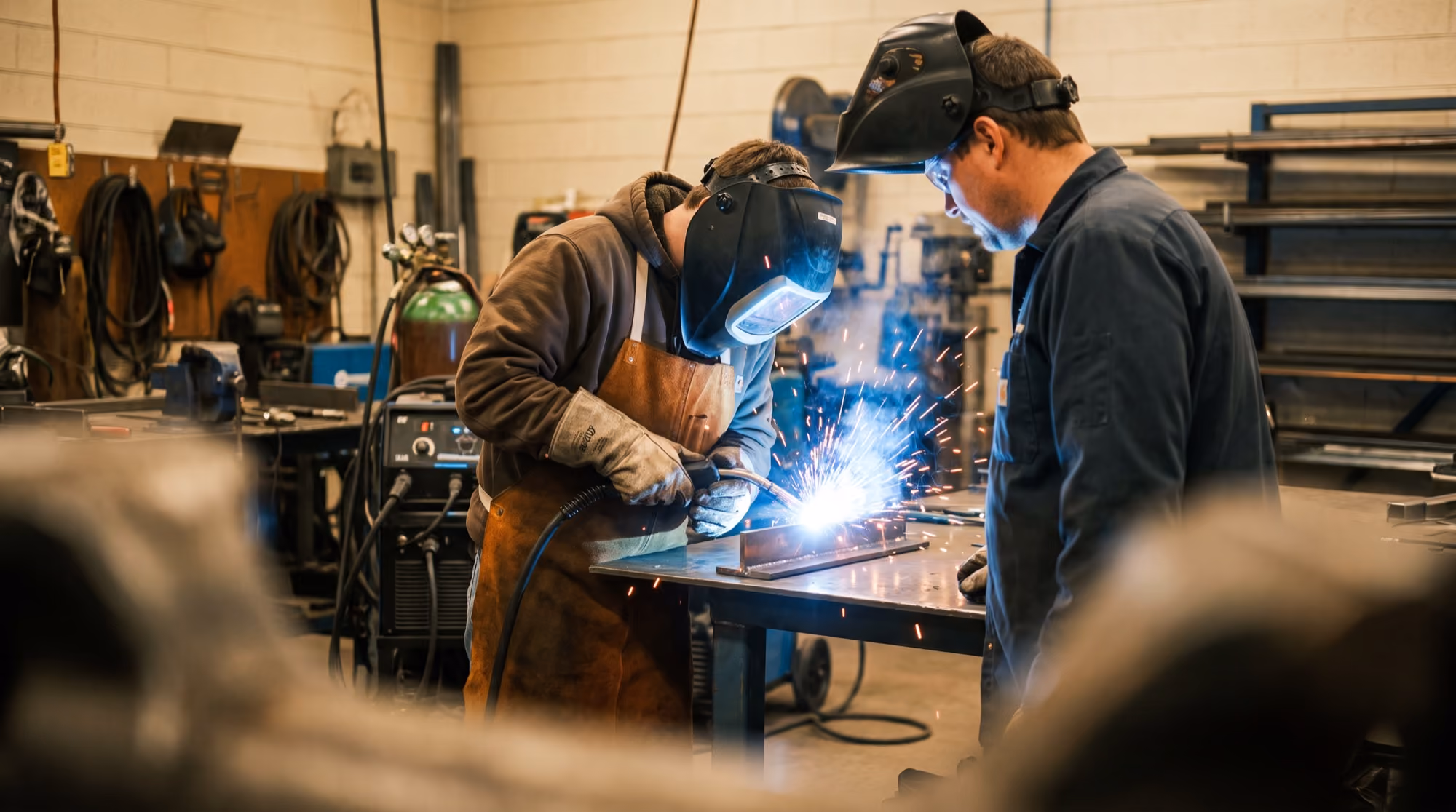 Welding instructor supervising a student mid-weld in a vocational trade-school shop