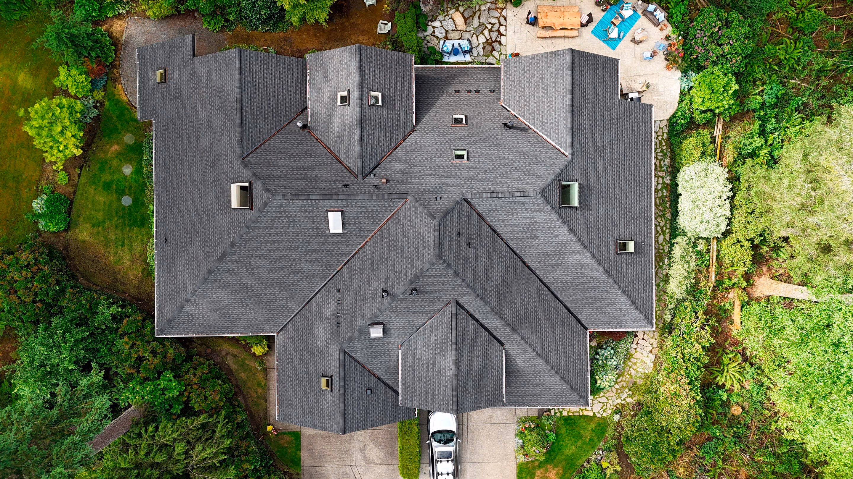 Aerial view of a large house with a dark gray complex roof surrounded by green trees and garden.