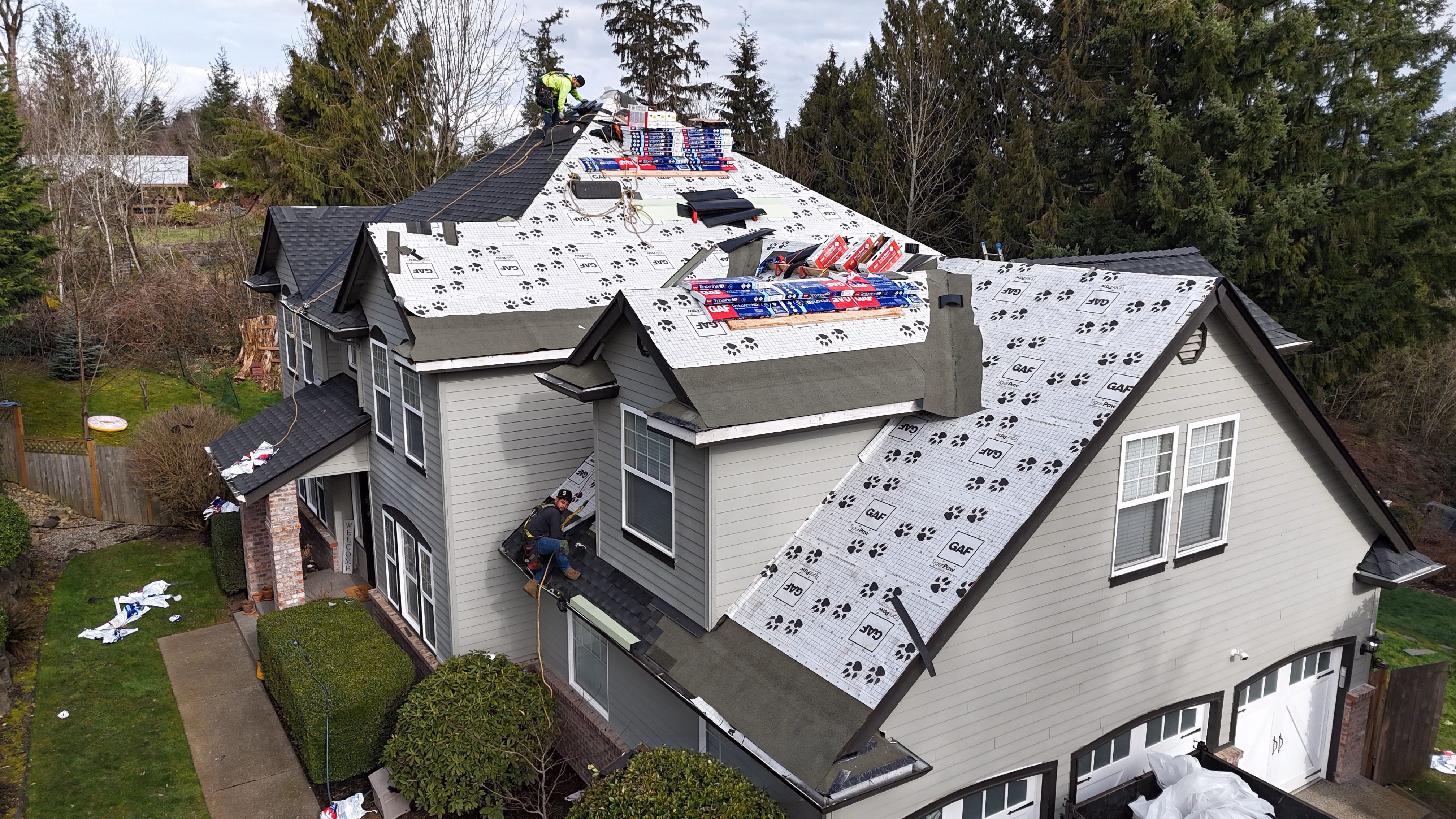 Two workers installing new roofing underlayment and shingles on a large house roof.
