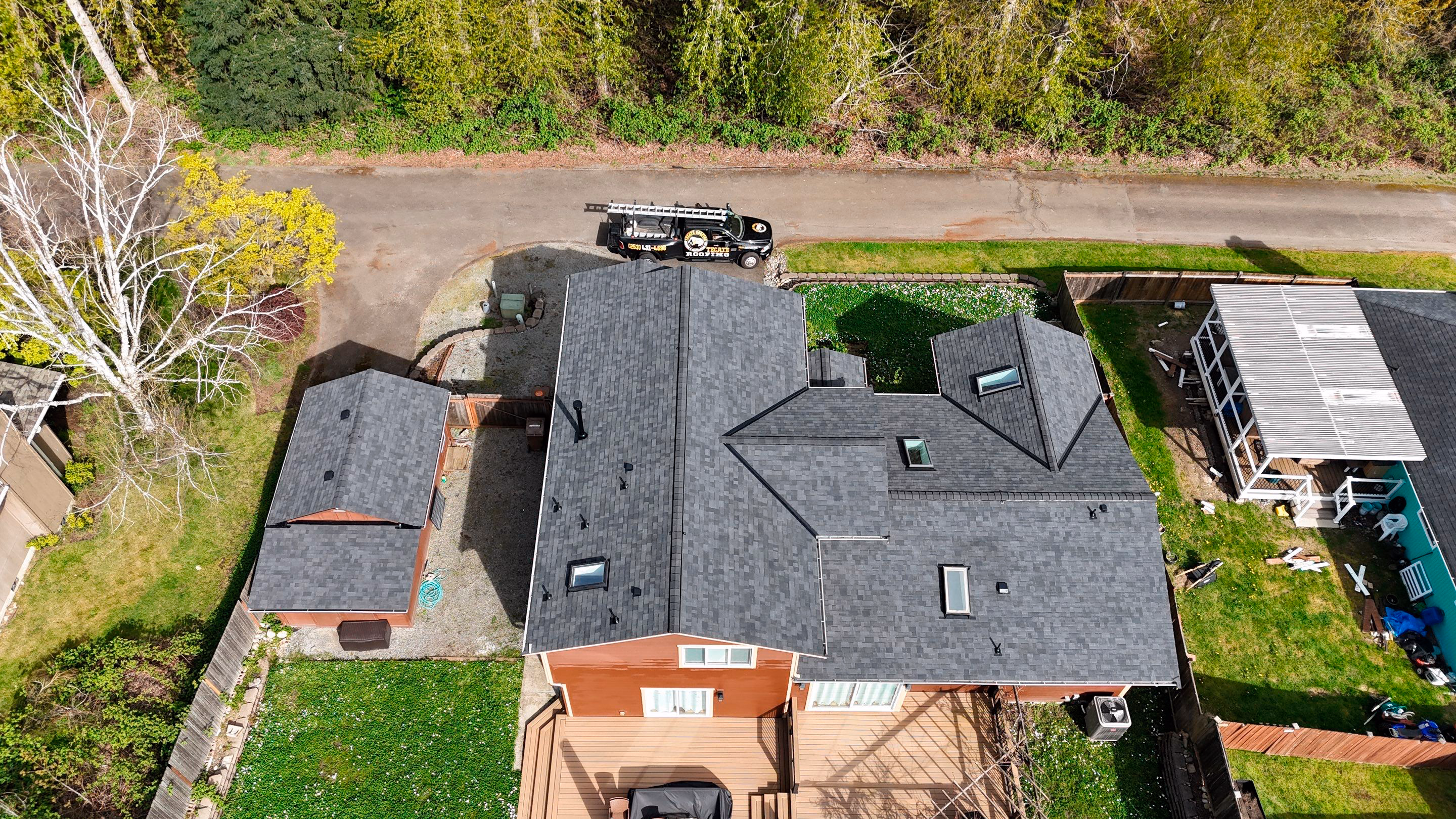 Aerial view of a house and a small shed with fresh dark gray roofing shingles on a sunny day.