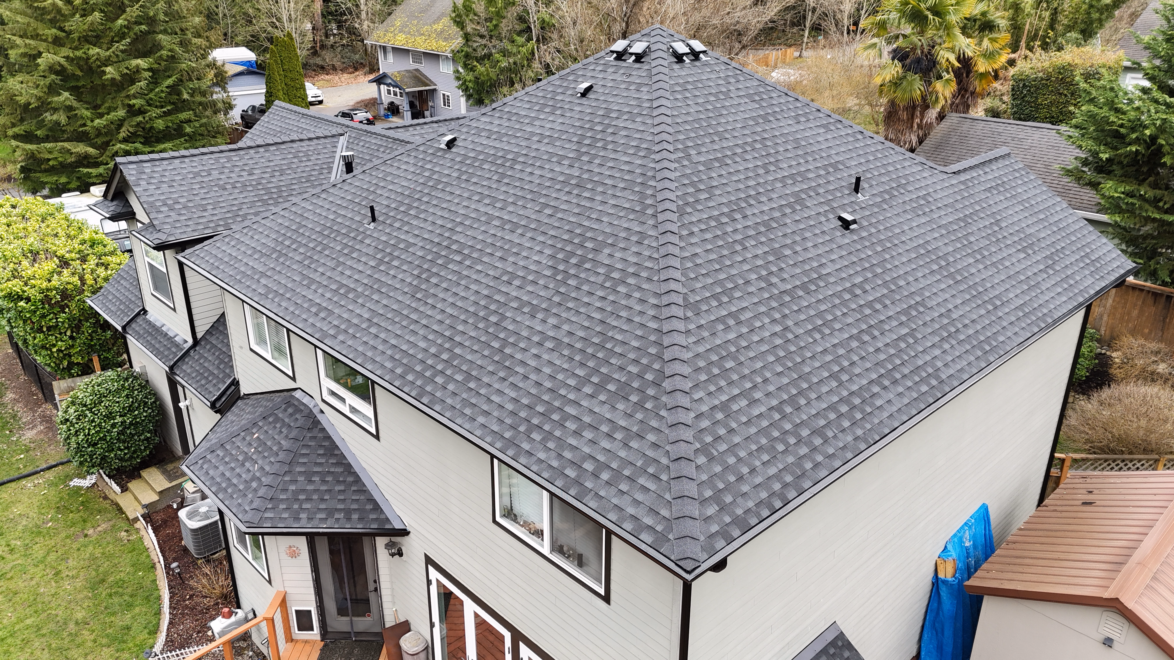 Aerial view of a large house with a newly shingled gray roof surrounded by trees and neighboring homes.