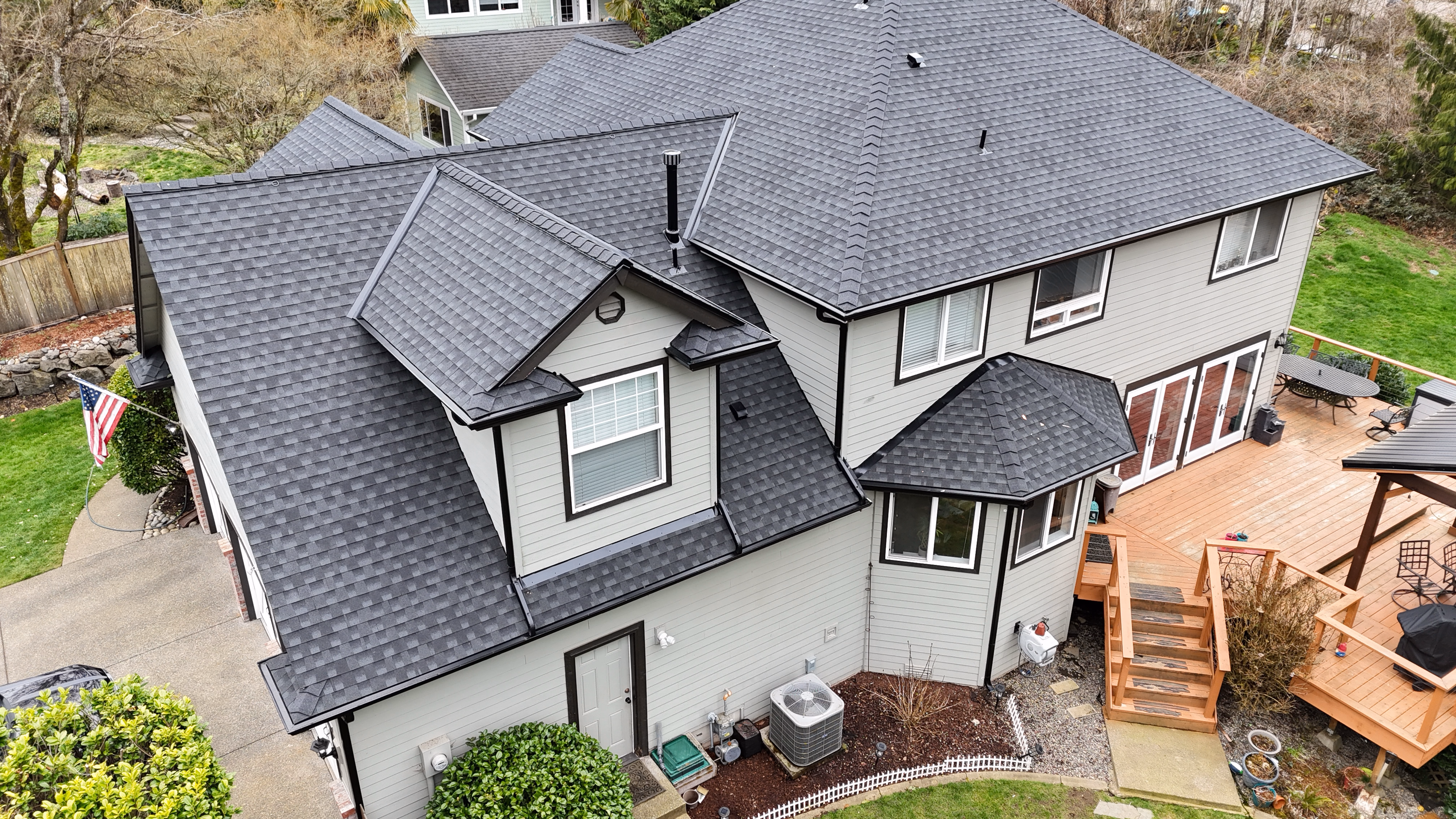 Aerial view of a large house with dark gray shingle roof and beige siding near wooden deck.