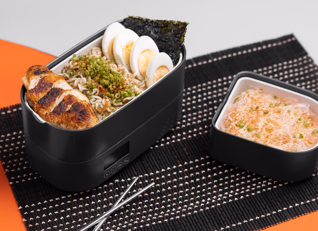 Black electric lunch box with grilled chicken, ramen noodles, sliced boiled eggs, seaweed, and side dish with rice.