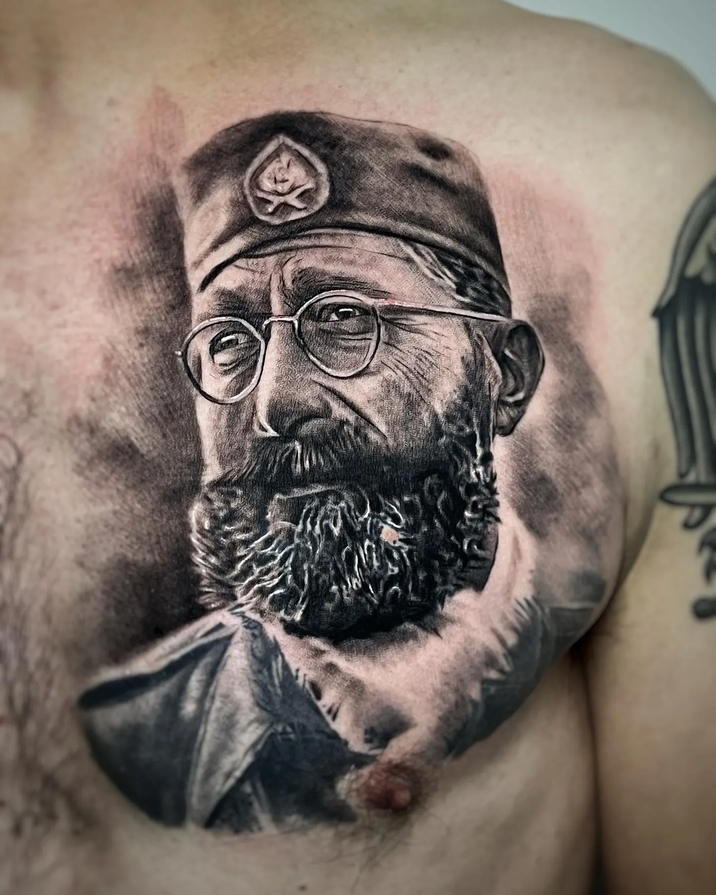 Realistisches Black and Grey Portrait Tattoo auf der Brust – Mann mit Vollbart, Nickelbrille und Militärmütze, INK SOCIETY