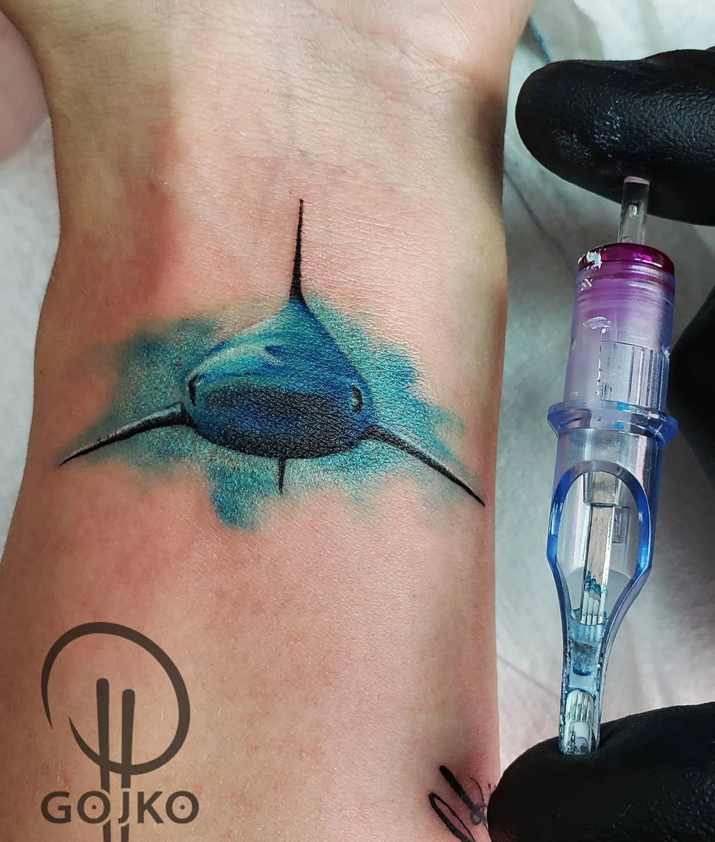 Farbiges Hai Tattoo am Handgelenk – realistischer Hai von vorne mit blauem Watercolor-Hintergrund, von Gojko, INK SOCIETY