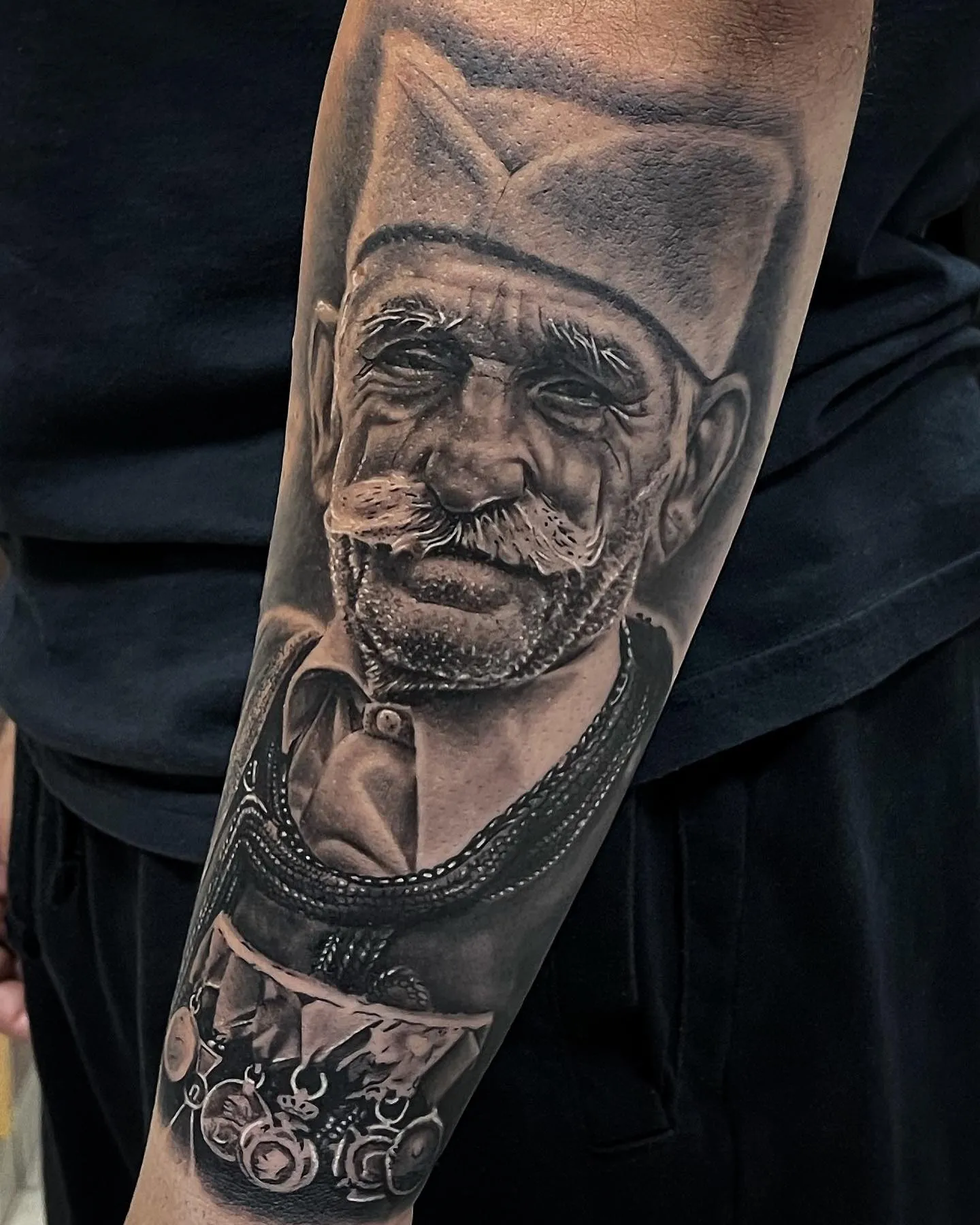 Realistisches Black and Grey Portrait Tattoo am Unterarm – älterer Mann mit Schnurrbart, traditioneller Mütze und Trachtenweste mit Orden, INK SOCIETY