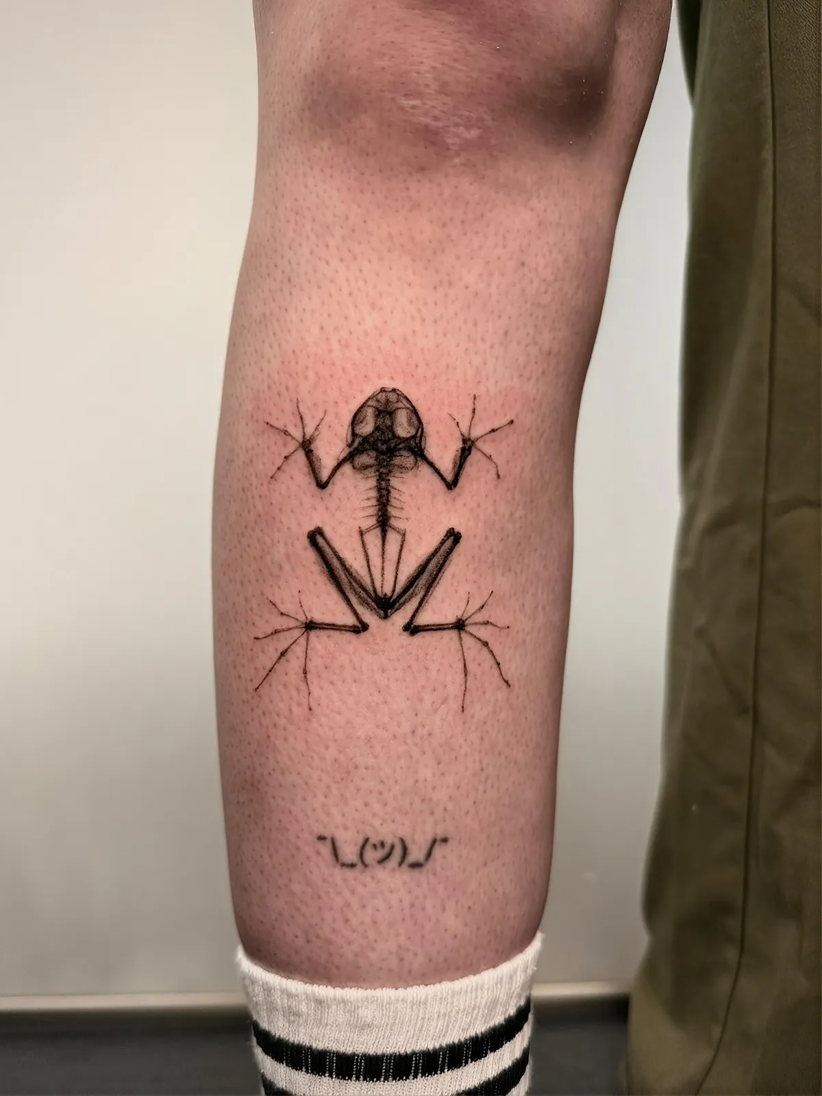 Frosch Skelett Tattoo am Unterschenkel – Froschskelett im Röntgen-Stil in Black and Grey, darunter ein kleines Shrug-Emoticon, von Lara, INK SOCIETY