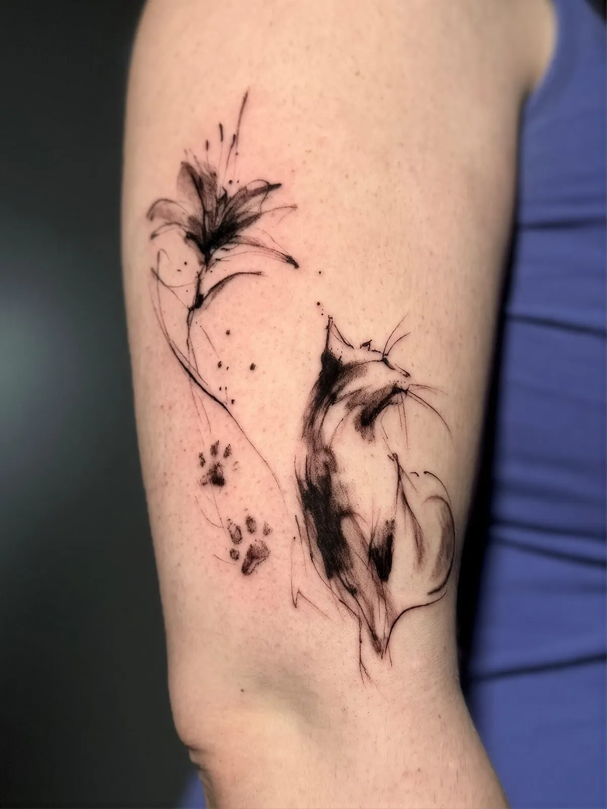 Katzen Tattoo am Oberarm im Sketch-Stil – sitzende Katze mit Lilie und Pfotenabdrücken in schwarzer Tusche, von Lara, INK SOCIETY