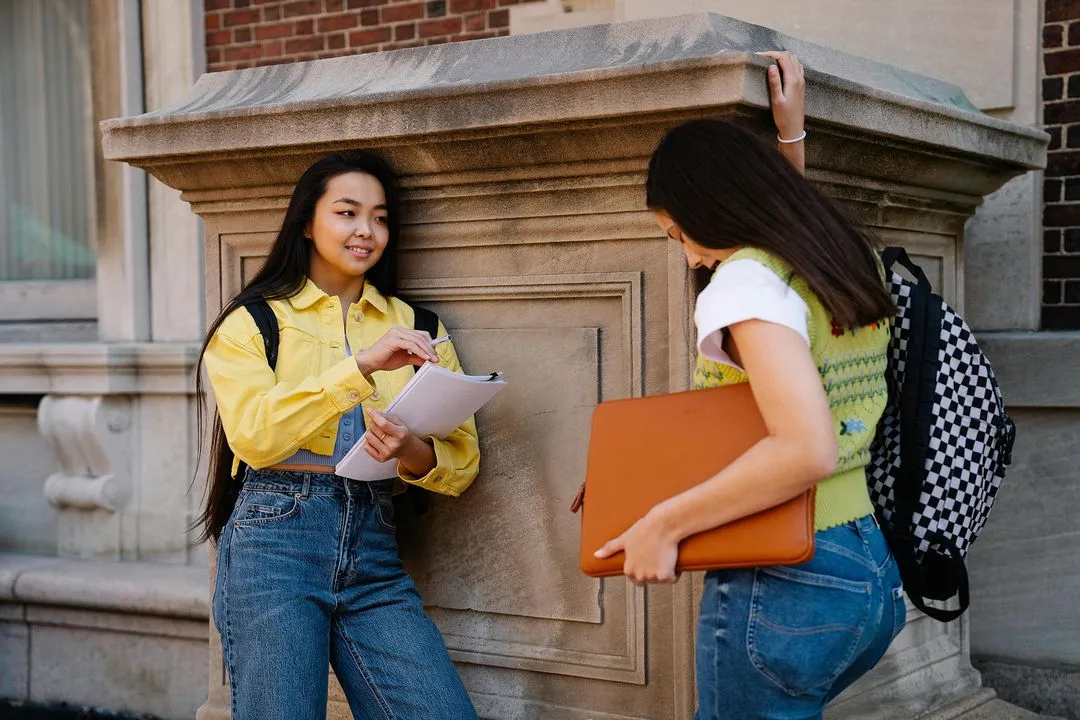 Two schoolgirls holding books are talking near a school building