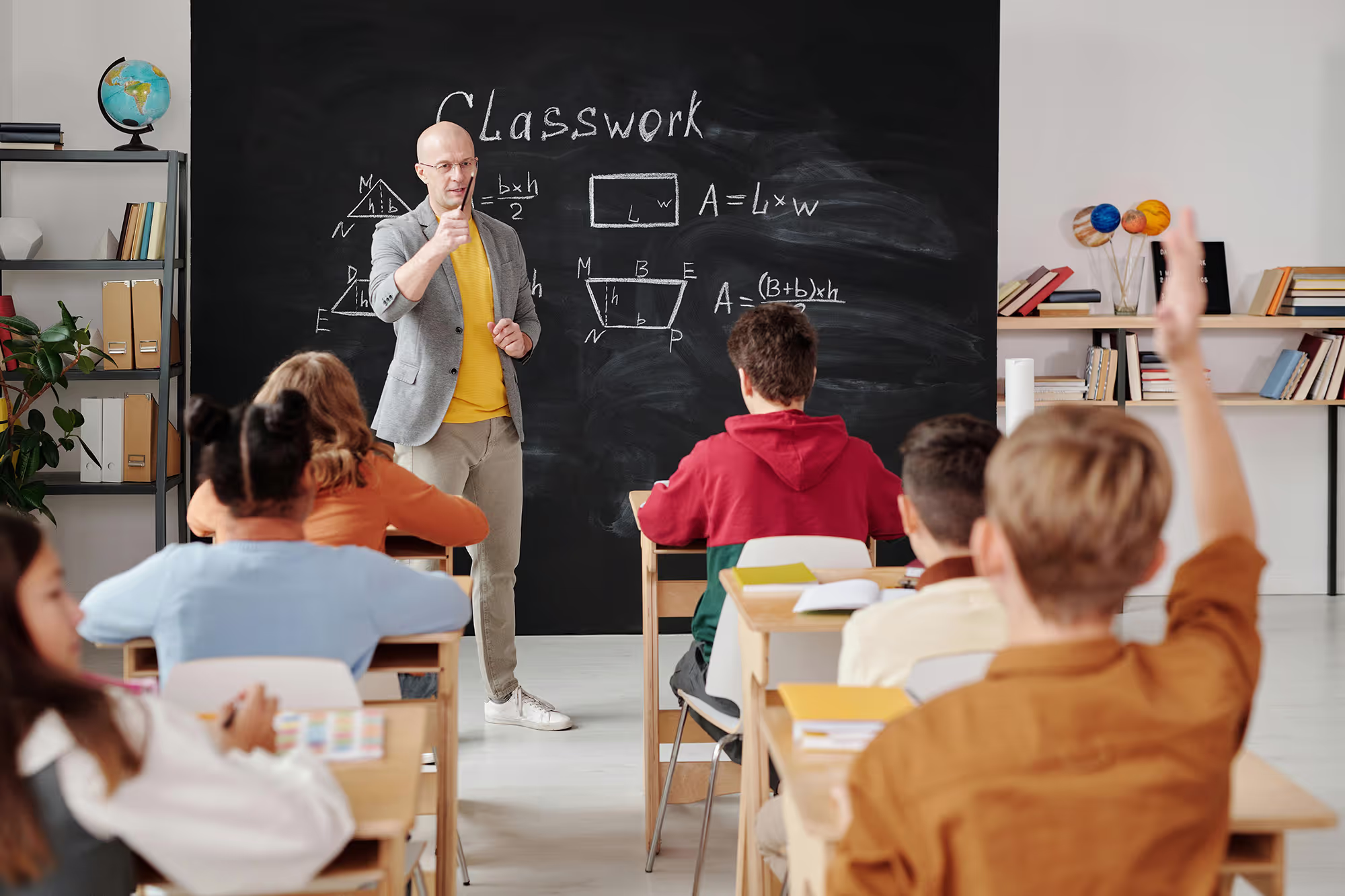 A teacher stands near a whiteboard in a classroom, explaining a lesson to the students