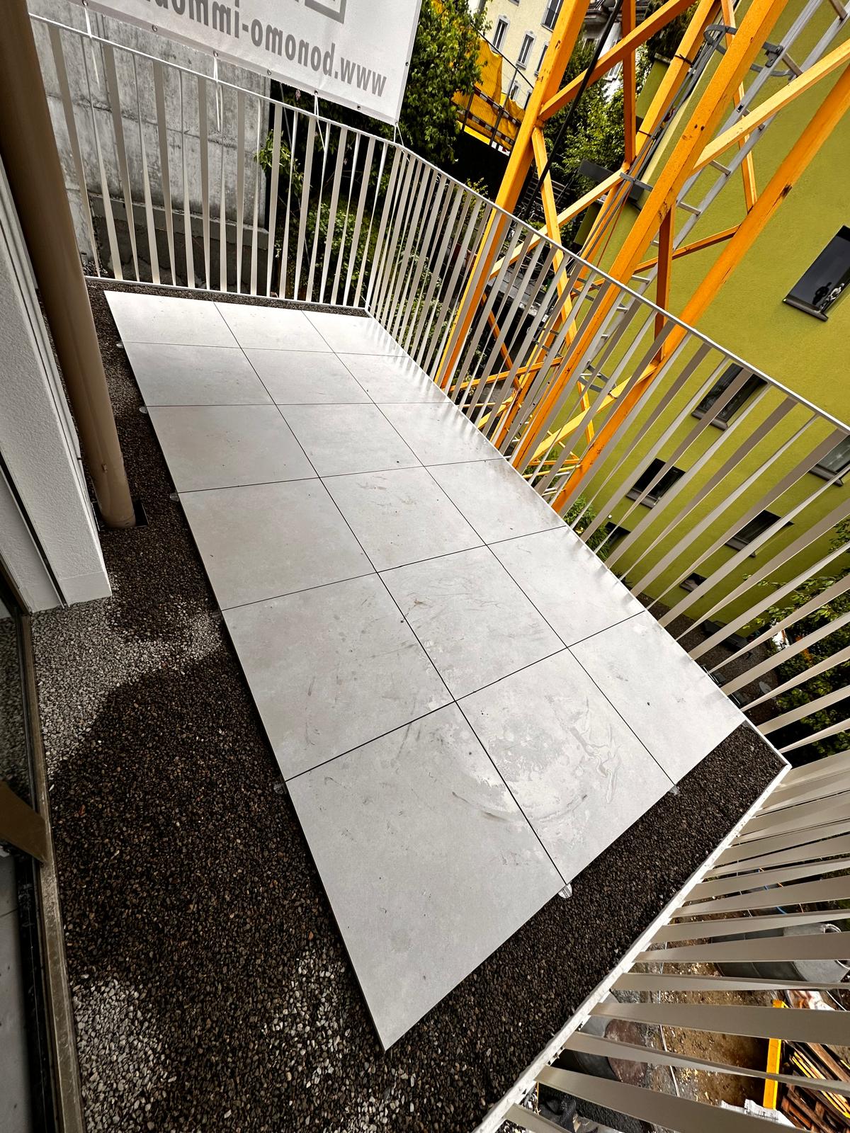 Balcony floor with large white square tiles installed over dark gravel, surrounded by white metal railing.