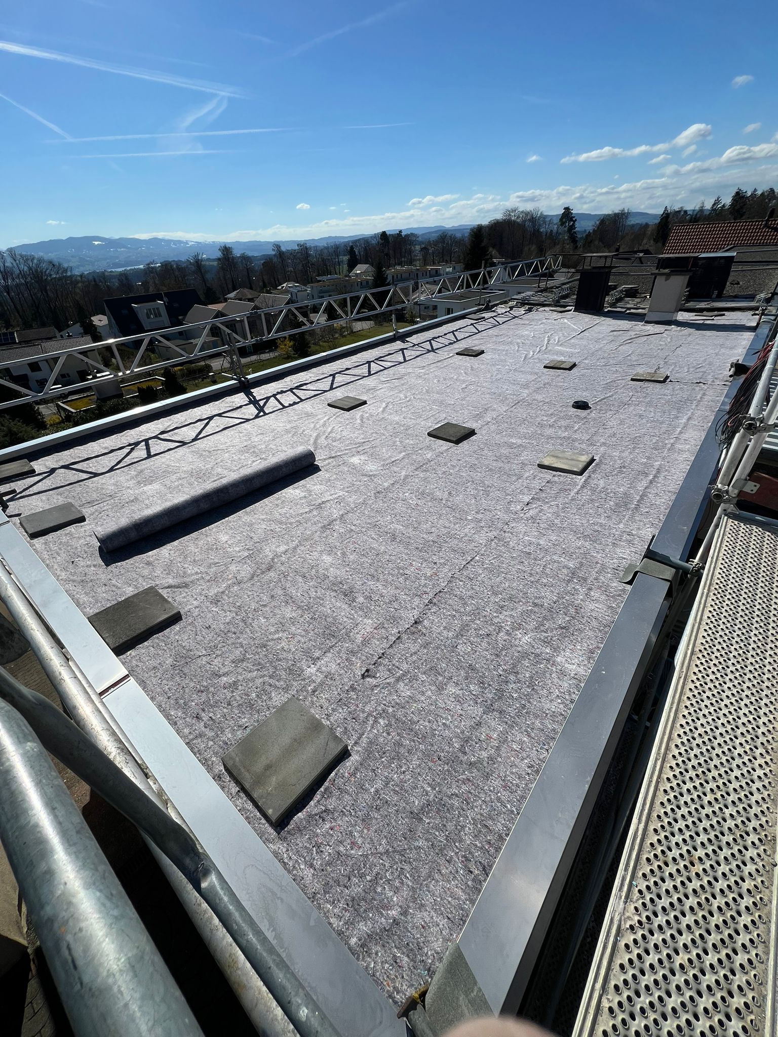 Flachdach mit einer Graufolie und Betonplatten, umgeben von Geländer, bei klarem Himmel.