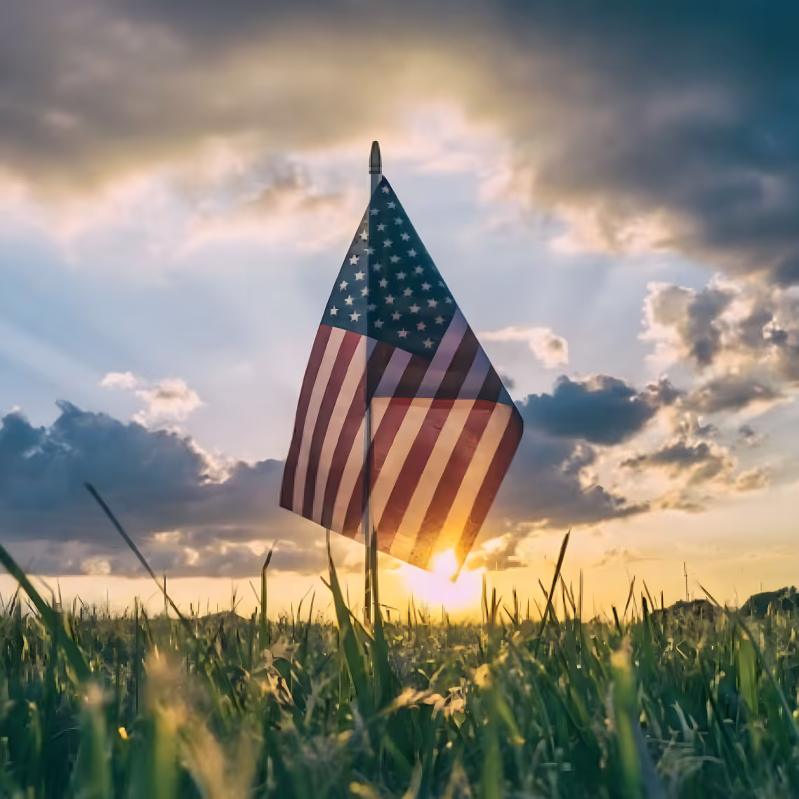 American flag on a pole in a grassy field at sunset with clouds and sun rays in the sky.