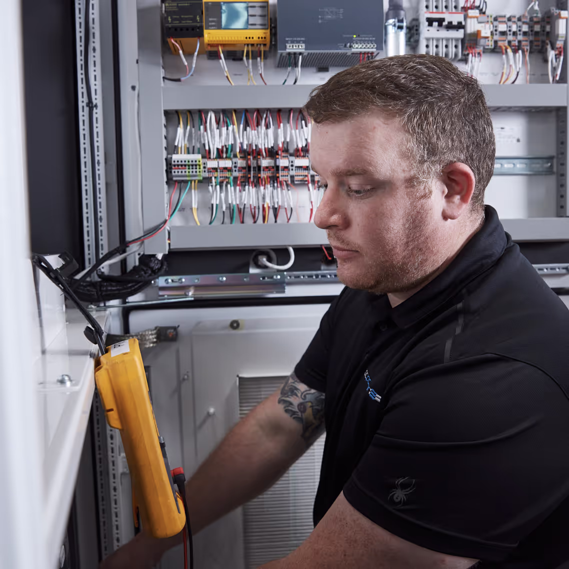Technician using a multimeter to test an industrial electrical control panel with multiple wires.