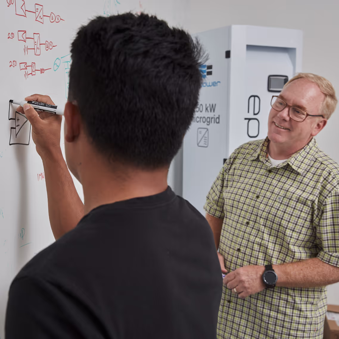 Man writes electrical diagrams on whiteboard while another man in plaid shirt watches smiling.