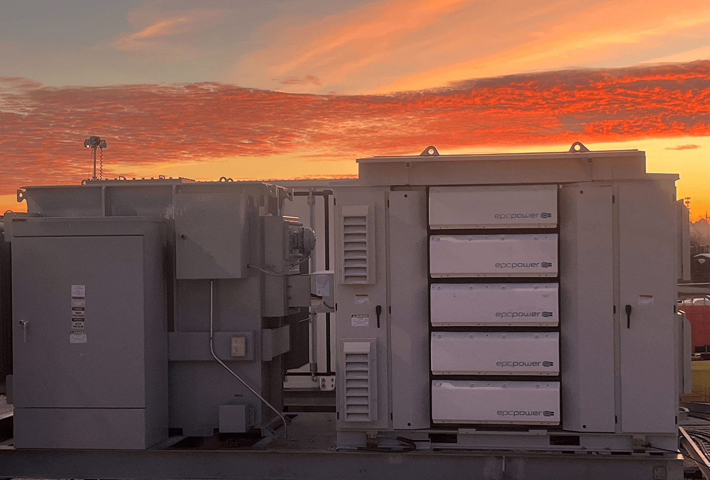 EPC Power M System unit on a rooftop at sunset, front face showing five stacked inverter modules against a vivid orange sky.