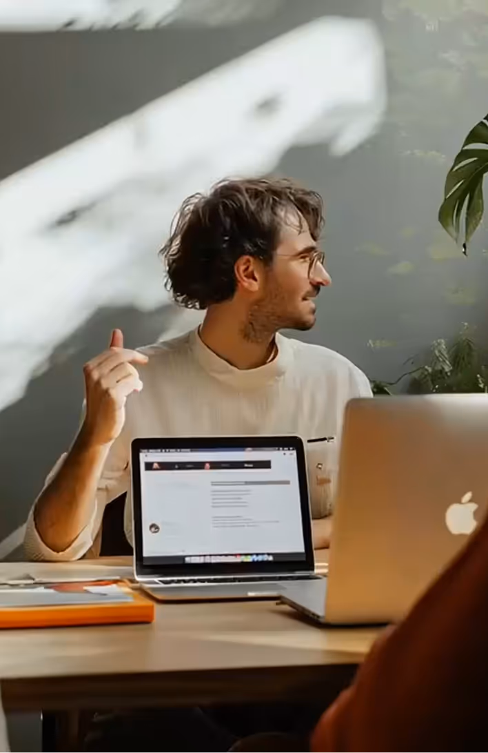 Man with curly hair and glasses sitting at a wooden desk with two open Apple laptops, gesturing with his hand.