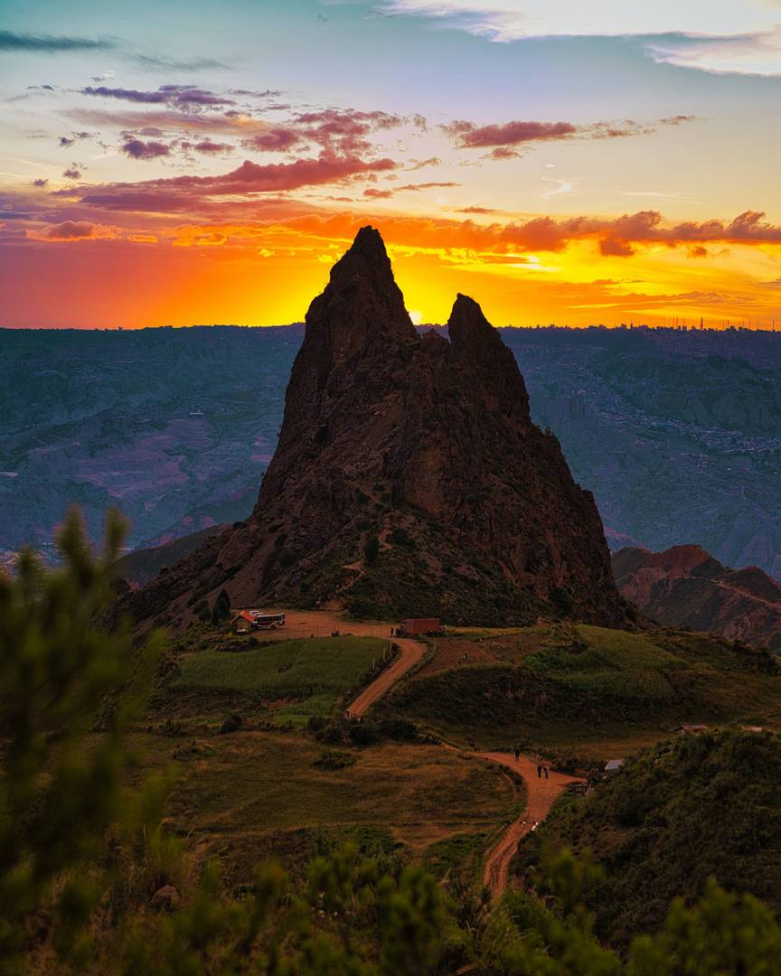 Tall rocky peak silhouetted against a vibrant orange and yellow sunset sky with scattered clouds, surrounded by green hills and a dirt road leading to a small building.