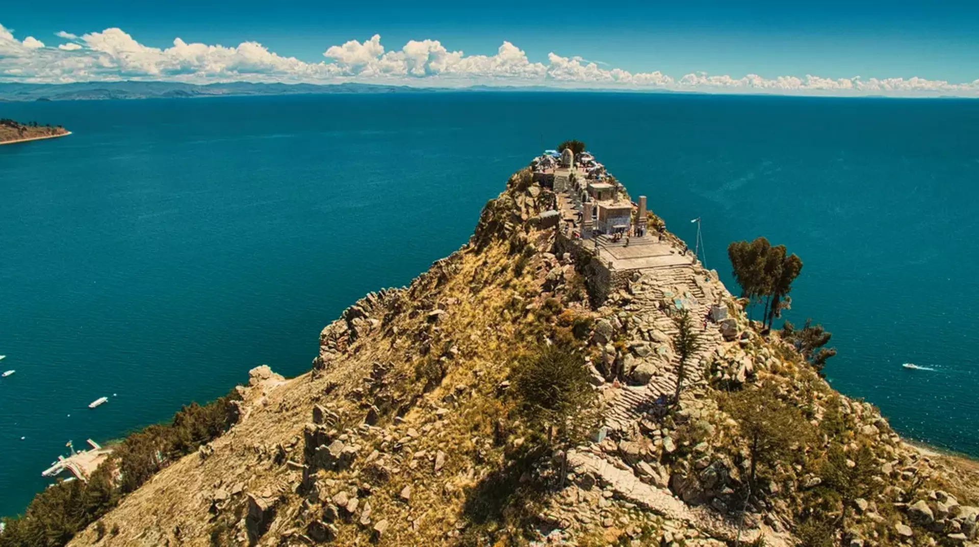 Panoramic view of a rocky hill with ancient stone terraces and pathways overlooking the deep blue waters of Lake Titicaca under a partly cloudy sky.
