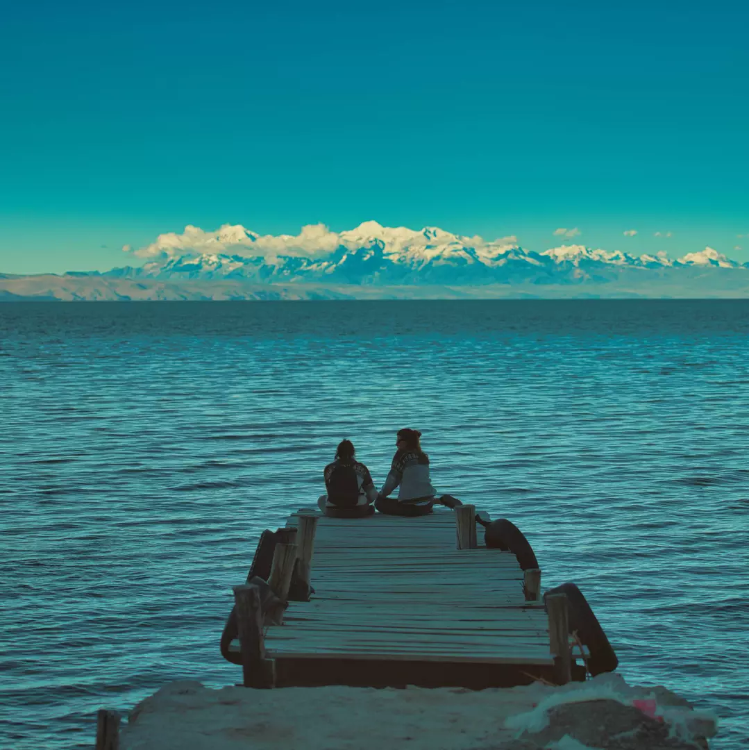 Two people sitting on the end of a wooden dock overlooking a large lake, with snow-capped mountains and a blue sky in the distance.