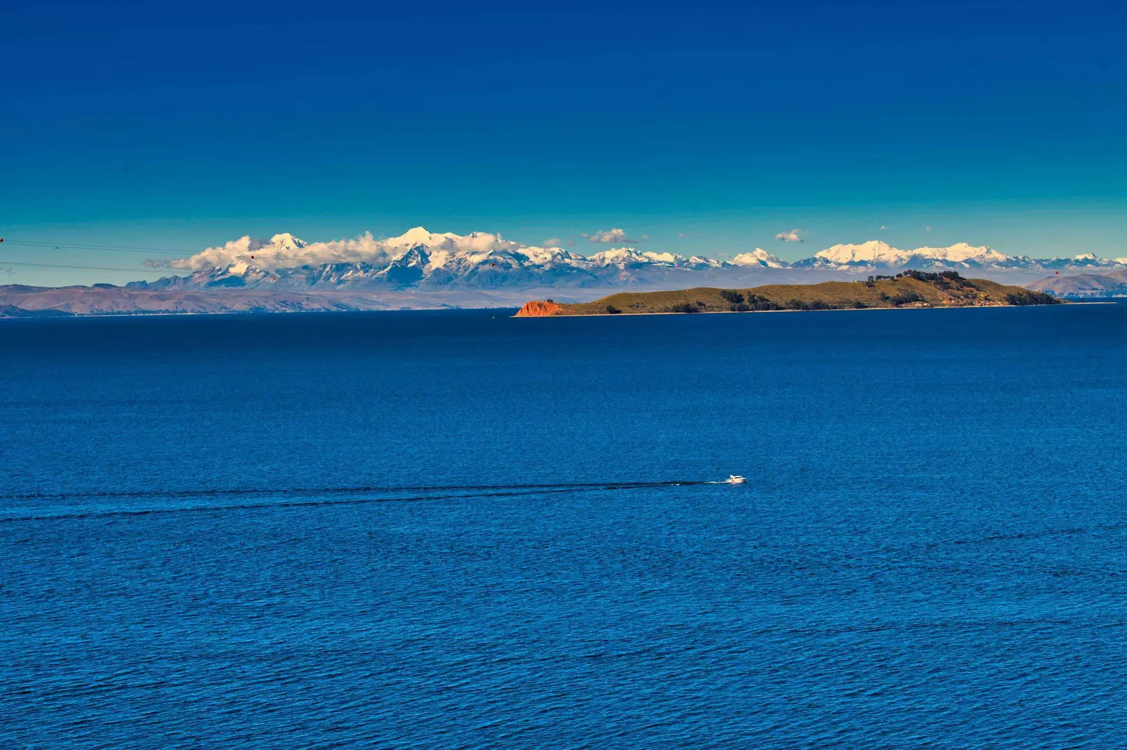Wide panoramic view of a deep blue lake with a small white boat creating a trail on the water, a green and brown island in the middle distance, and snow-capped mountains with clouds in the background under a clear blue sky.
