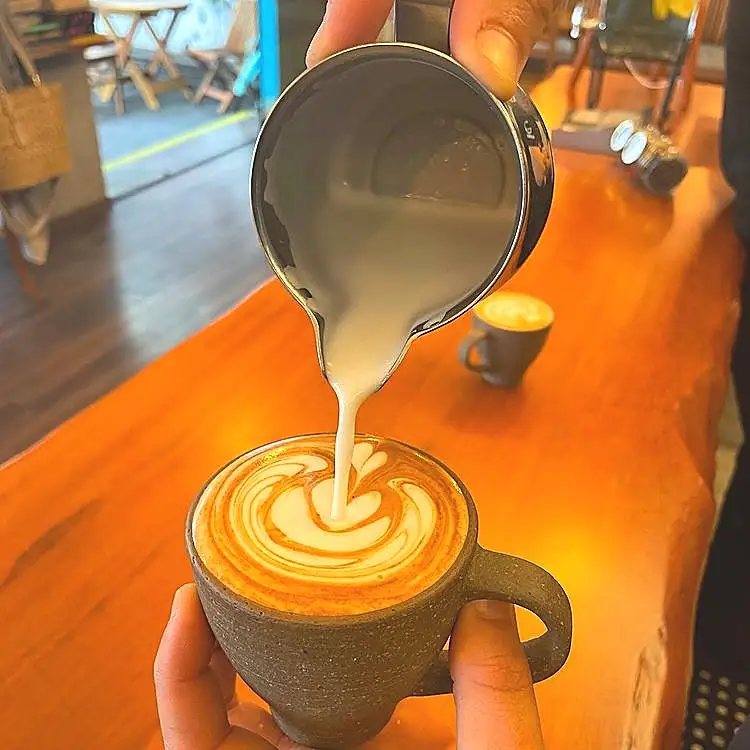 Person pouring steamed milk from a metal pitcher into a cup of coffee with latte art on top, on a wooden table in a cafe setting.
