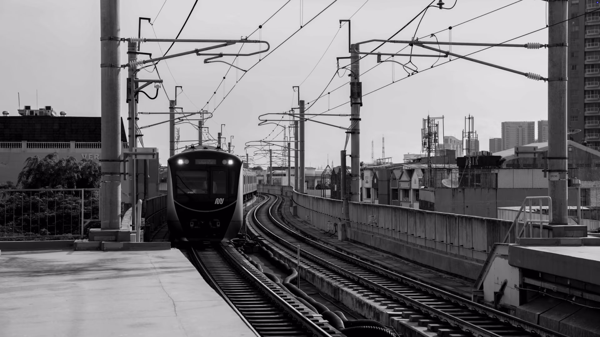 Black and white photo of a modern train approaching on elevated tracks in an urban area with buildings and electrical infrastructure around.