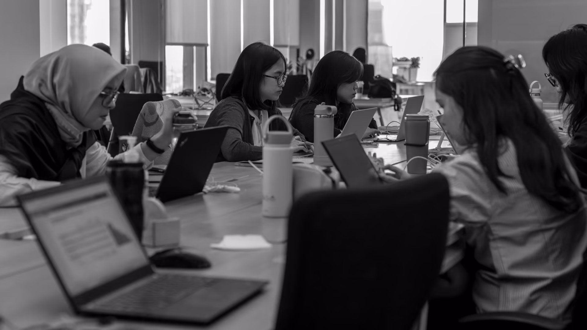 Black and white photo of five people sitting around a large table in an office, working on laptops with water bottles and coffee cups nearby.