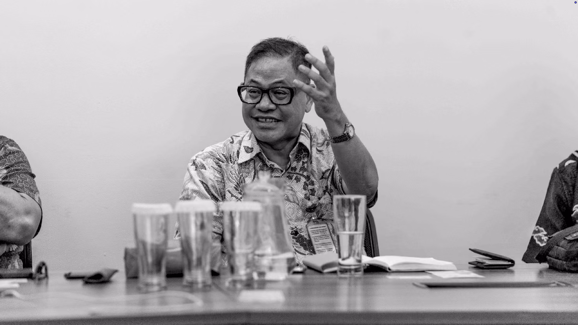 Smiling man wearing glasses and a patterned shirt gestures with his right hand while sitting at a table with several glasses, notebooks, and a water pitcher.