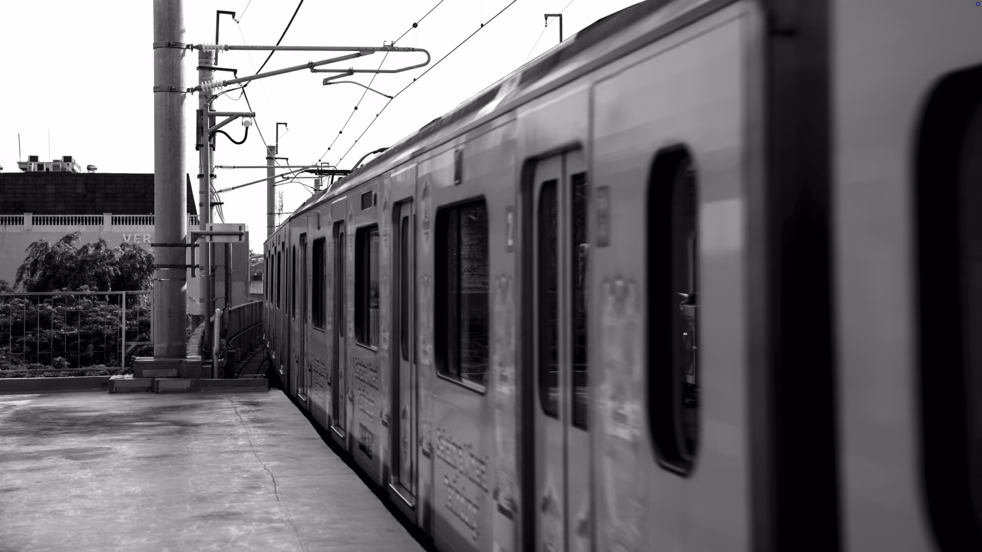Black and white photo of a commuter train stopped at an outdoor platform with overhead electrical wires visible.