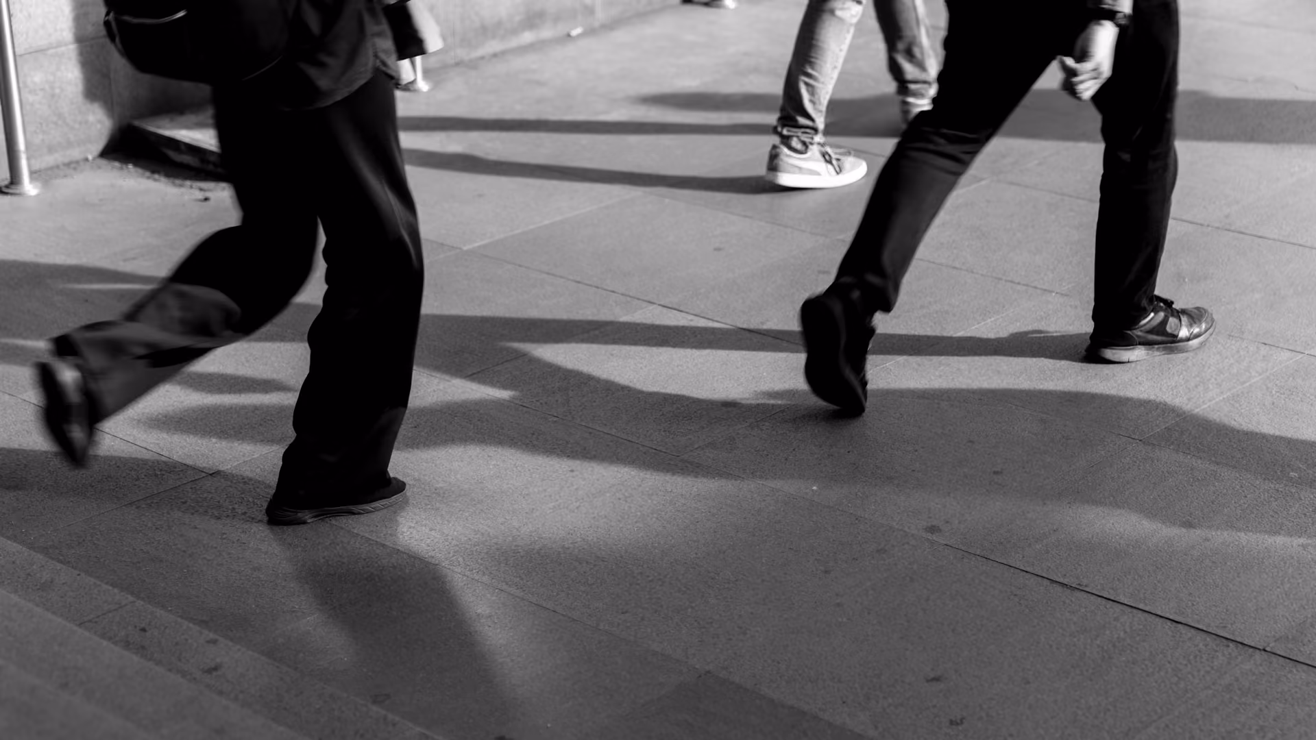Black and white photo of people walking on a sidewalk, showing only their legs and feet dressed in casual shoes and pants.