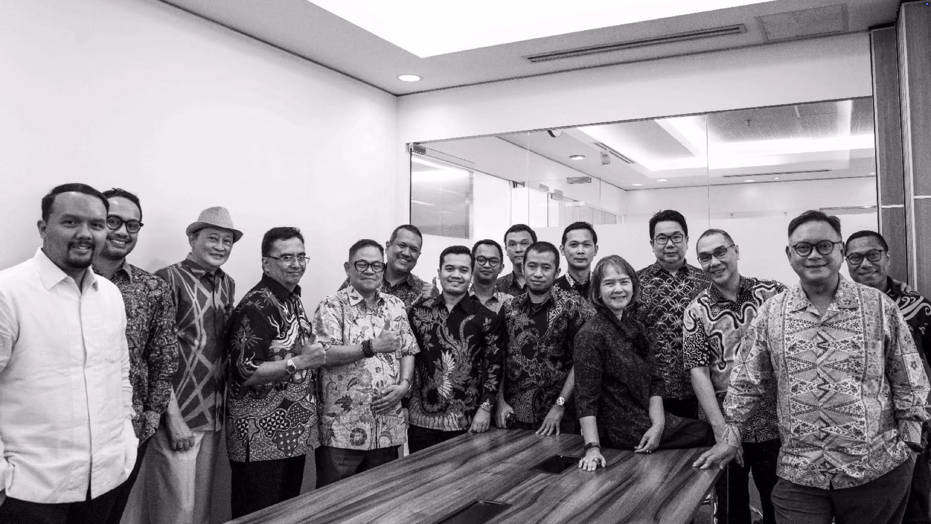 Group of sixteen people standing and smiling around a wooden conference table in a modern office room with a large glass wall in the background, all wearing patterned batik shirts except one person in a plain white shirt.