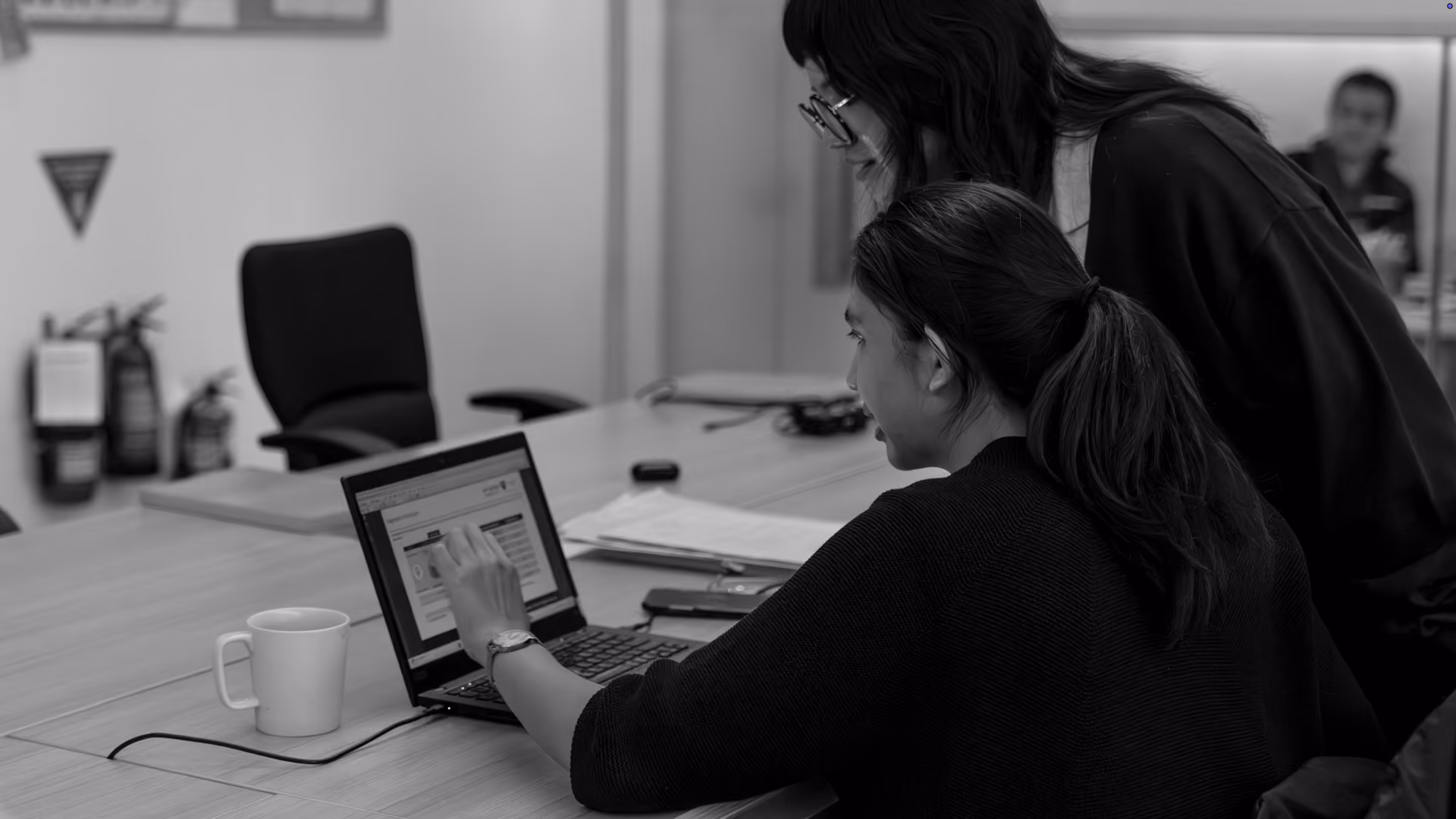 Two women working together at a desk with one pointing at a laptop screen, a coffee mug nearby, and papers on the table.