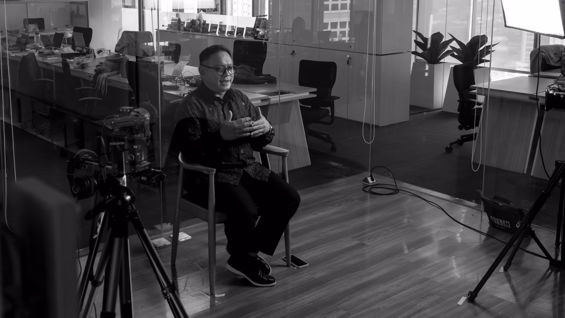 Man with glasses seated on a wooden chair speaking in an office, surrounded by filming equipment including cameras and lighting.